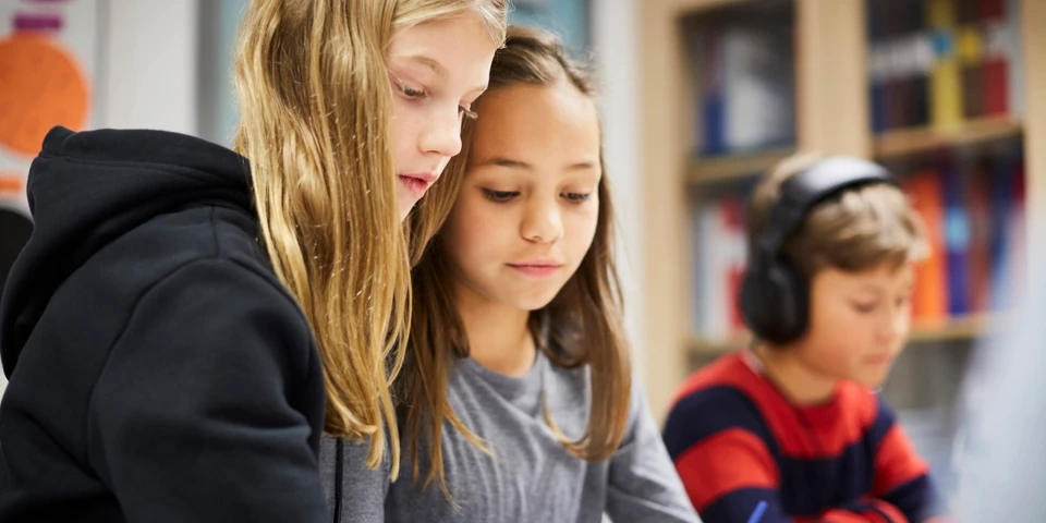 Tre elever. En har på sig hörlurar. Två tittar på något på bordet.